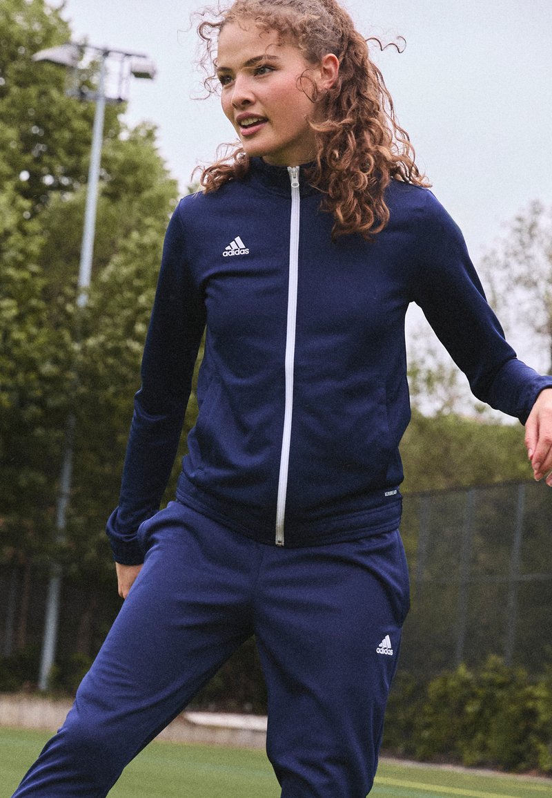 Marineblaues Sportanzug mit Reißverschlussjacke und elastischen Hosen. Verfügt über das weiße Adidas-Logo auf der oberen linken Seite und am Hosenbein. Glattes Material.
