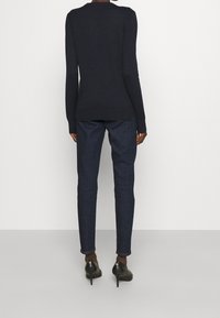 En person står med ryggen mot kameran, iklädd en svart långärmad tröja, mörkblå åtsittande byxor och svarta högklackade skor mot en neutral bakgrund.