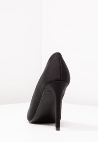 Chaussure noire à talon haut avec un bout pointu et une finition satinée élégante. Le talon est fin, de style stiletto, avec une silhouette lisse et courbée.