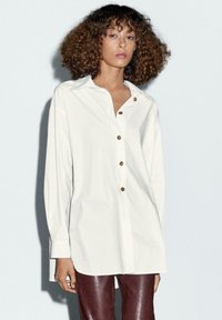 Camicia bianca con colletto classico, dotata di bottoni color oro. Maniche lunghe, vestibilità comoda, tessuto morbido e una sottile lucentezza.