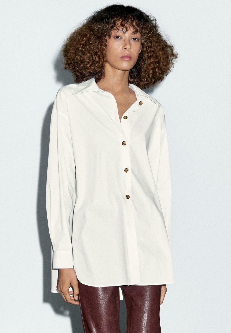 Camicia bianca con colletto classico, dotata di bottoni color oro. Maniche lunghe, vestibilità comoda, tessuto morbido e una sottile lucentezza.
