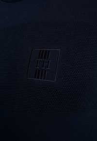 Granatowy dzianinowy wyrób odzieżowy z teksturowanym wzorem i wypukłym logo "TH" na kwadratowej naszywce, z okrągłym dekoltem i elastycznym materiałem.