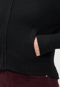 Svart stickad tröja med en texturerad yta, med dragkedja fram, ribbade mudd och sidofickor. Bärs över bordeauxfärgade kordbyxor.