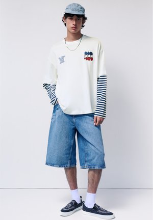 Jeune homme portant une casquette bleue, une chemise blanche à manches longues superposée avec des rayures, un short en jean longueur genou, des chaussettes blanches et des chaussures bateau bleu marine.