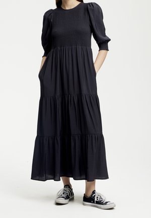 Femme portant une robe midi noire à étages avec des manches bouffantes et des baskets noires basses, debout avec les mains dans les poches de la robe.