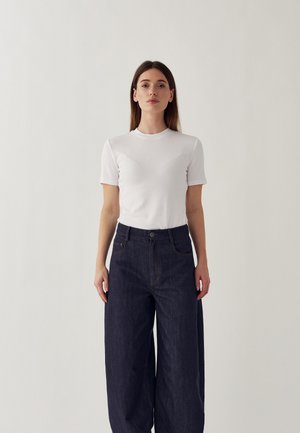 Biele tričko s krátkym rukávom a priliehavým strihom, v kombinácii s voľnými tmavomodrými džínsovými nohavicami s prednými vreckami a zapínaním na gombík.