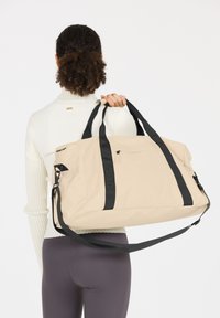 Bolsa de lona beige con asas negras y correa ajustable; cuenta con un bolsillo frontal y una tela suave y texturizada.