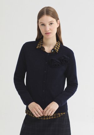 Jeune femme aux cheveux bruns et raides porte un cardigan noir avec des détails floraux sur une chemise à carreaux marron et noire.