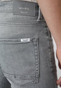 Jeans in denim grigio con un taglio slim. Presenta un'etichetta con la dicitura "Marc O'Polo DENIM" e particolari di cucitura sulla tasca posteriore. La texture appare liscia.