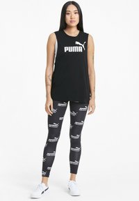 Svart ärmlös tröja med vitt "PUMA"-logotyp, parat med leggings som har ett upprepande svart och vitt PUMA-mönster, vita sneakers.