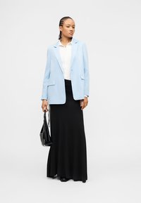 Γυναίκα με πλεξούδες, που φοράει γαλάζιο σακάκι, λευκό πουκάμισο, μακριά μαύρη φούστα, κρατώντας μαύρη τσάντα, στέκεται μπροστά σε απλό φόντο.