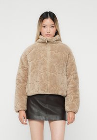 Kvinde iført beige fuzzy zip-up jakke og sort læder mini nederdel, stående imod en ensfarvet lys baggrund, vender fremad.