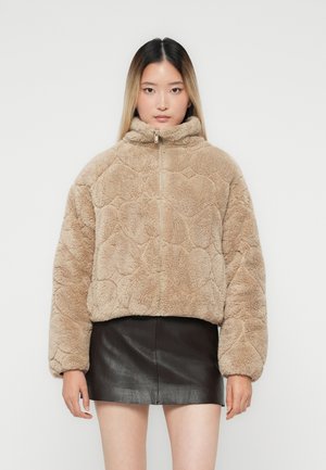 Kvinna i beige fluffig zip-up jacka och svart läder minikjol, står mot en enkel ljus bakgrund, vänd framåt.