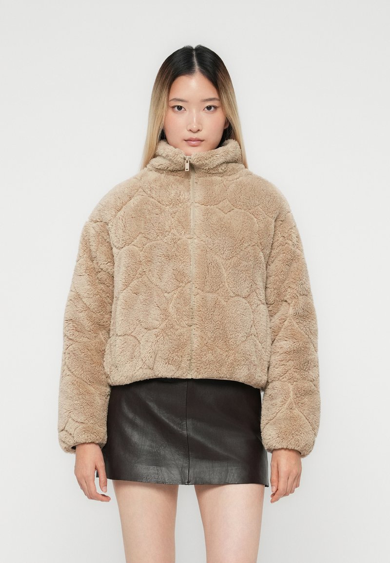 Kvinde iført beige fuzzy zip-up jakke og sort læder mini nederdel, stående imod en ensfarvet lys baggrund, vender fremad.