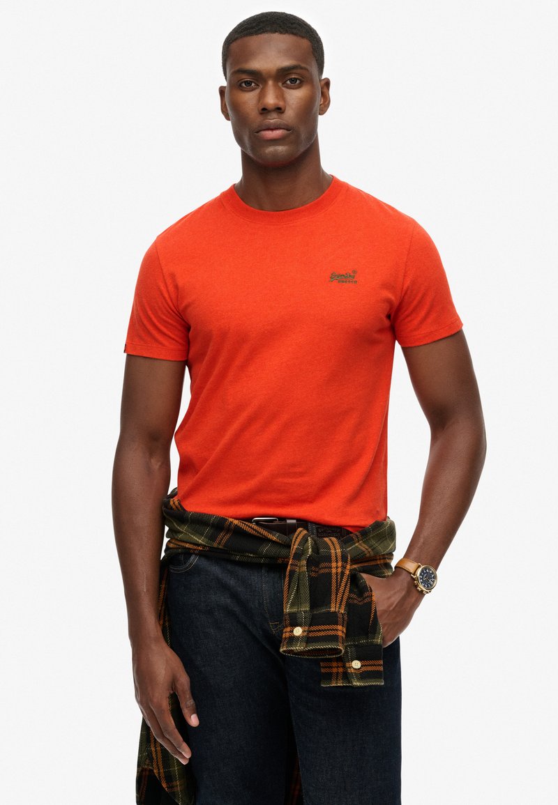 Man in een feloranje t-shirt, donkere spijkerbroek, een groen en oranje geruite overhemd om zijn taille geknoopt, en een gouden horloge om zijn linkerpols.