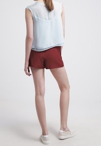 Vaalea sininen hihaton pusero, jossa on läpinäkyvä selkä ja yksityiskohtaiset olkaimet, yhdistettynä maroonivärisiin shortseihin ja beigeihin liukuhyppykenkiin.