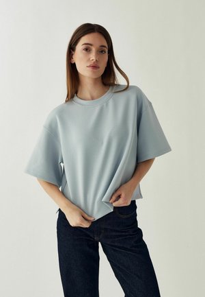 Vrouw met steil bruin haar, gekleed in een losse lichtblauwe korte mouwen blouse en donkere spijkerbroek, houdt de zoom van het shirt met één hand vast.