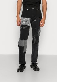 Hombre que lleva jeans de patchwork negros con parches de tela gris y estampados, y zapatos de cuero negros, de pie contra un fondo blanco.