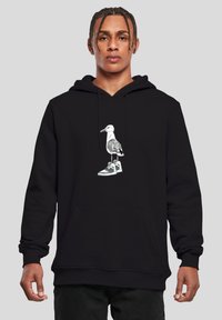 Jeune homme portant un sweat à capuche noir avec un graphisme d'une mouette debout sur une sneaker, sur un fond gris uni.