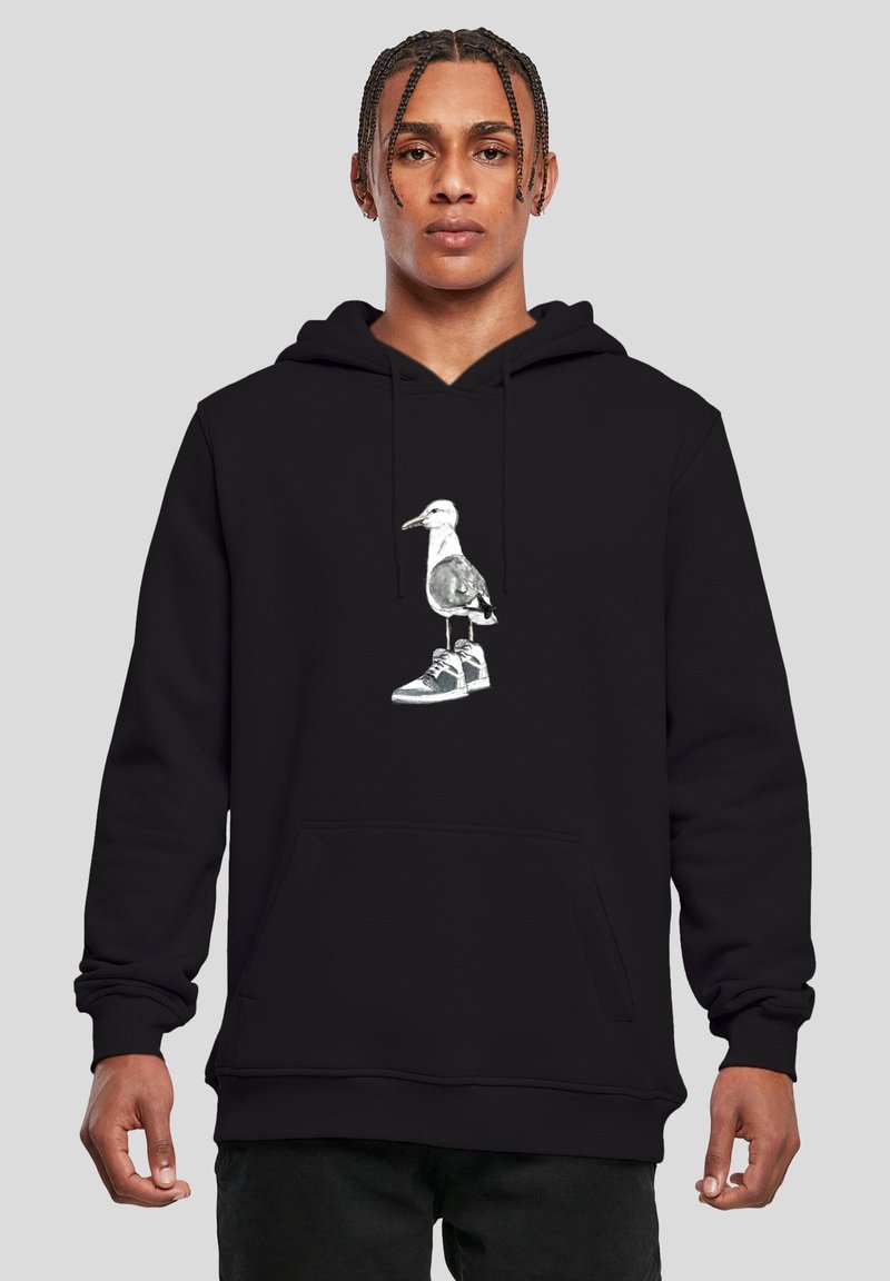 Jeune homme portant un sweat à capuche noir avec un graphisme d'une mouette debout sur une sneaker, sur un fond gris uni.