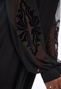 Schwarze, durchsichtige Stoffärmel mit aufwendigem, samtartigem Design, getragen über dunkelhäutiger Haut, am Handgelenk mit schwarzem, festem Stoff gerafft.