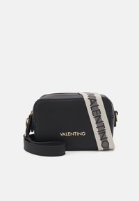 Bolso cruzado de cuero negro con un logo dorado, forma rectangular y una correa gris que muestra el nombre de la marca. Cierre superior con cremallera.