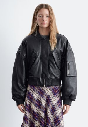 Donna con lunghi capelli biondi che indossa una giacca di pelle nera oversize e una gonna a quadri blu, bianca e rossa, in piedi davanti a uno sfondo semplice.