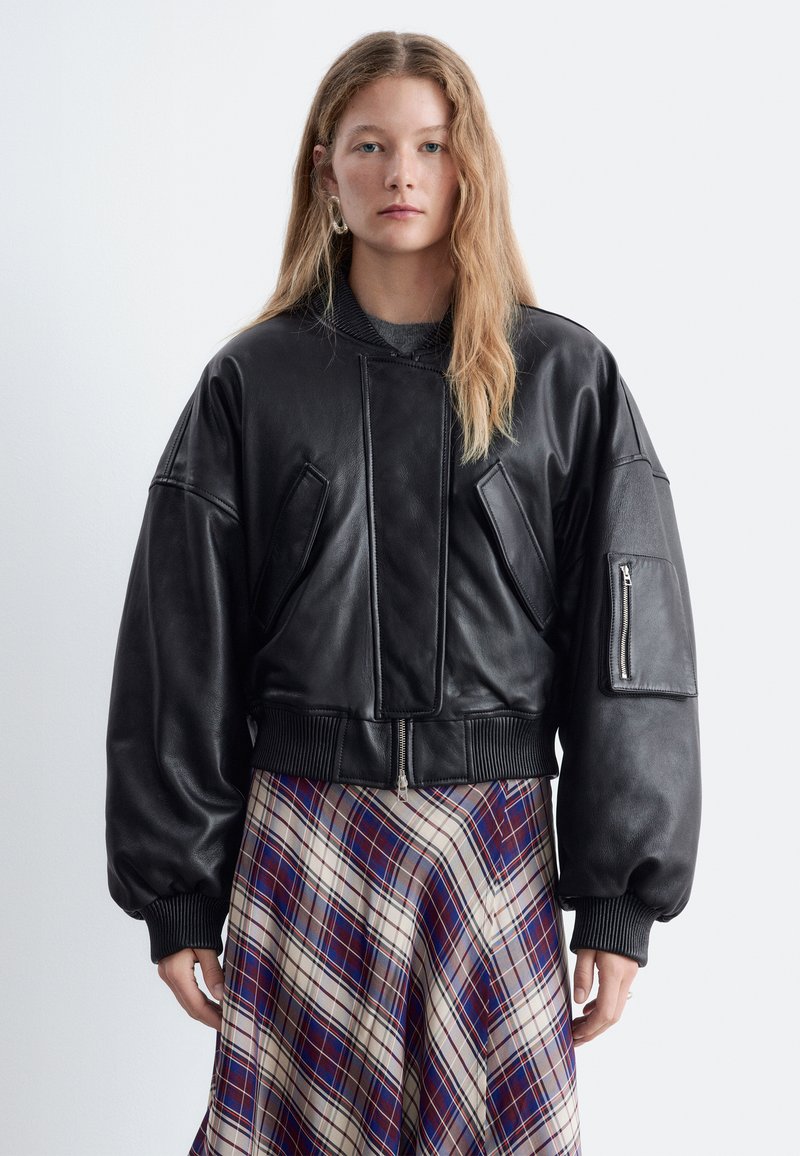 Femme aux longs cheveux blonds portant une veste en cuir noire oversize et une jupe à carreaux bleue, blanche et rouge, debout devant un fond uni.