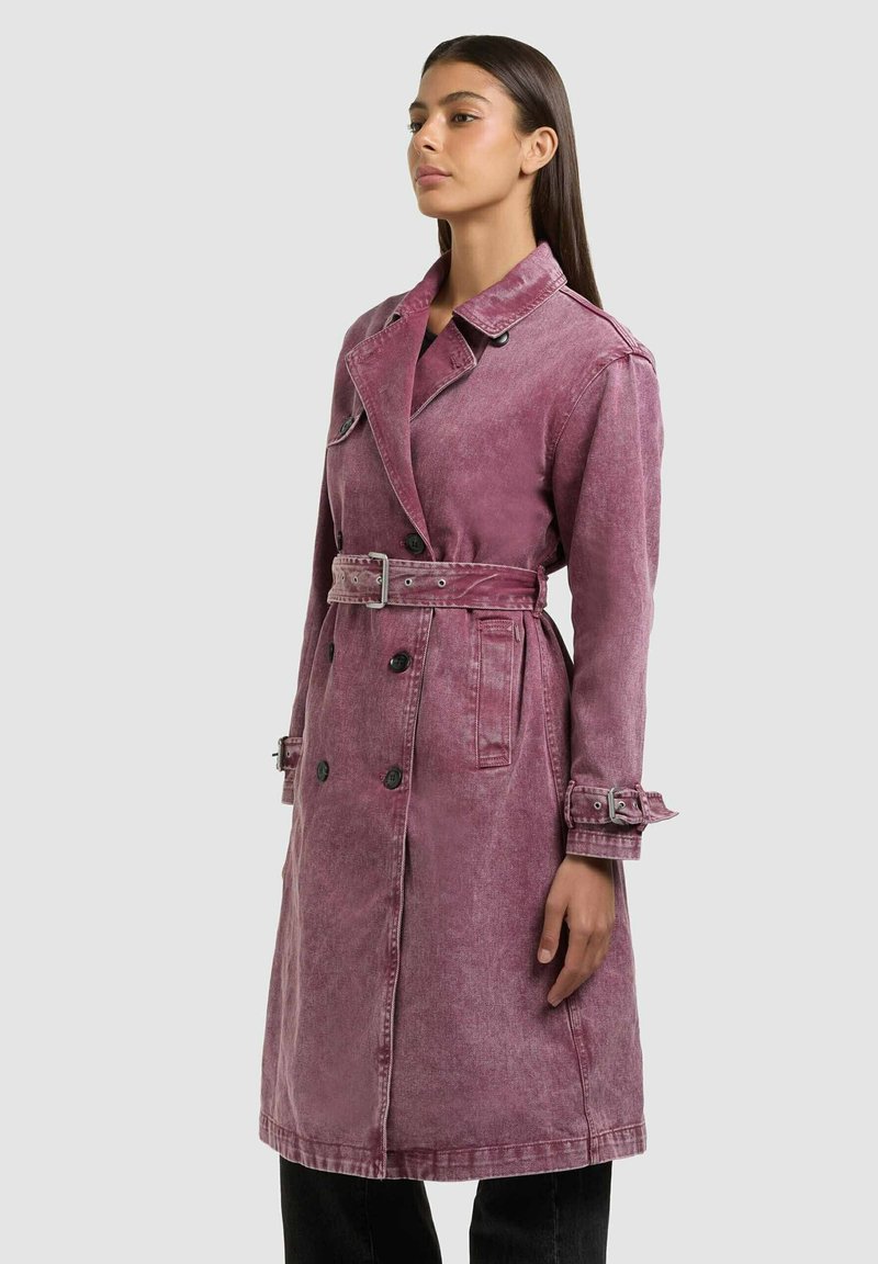 Donna con capelli lunghi e scuri che indossa un trench rosa polvere doppiopetto con cintura, bottoni neri e cinturini ai polsi, in piedi di profilo.