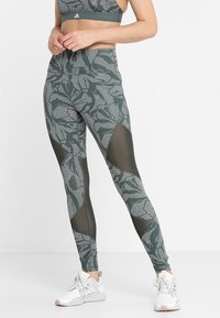 Kobieta w zielono-białych legginsach w wzory z czarnymi siateczkowymi wstawkami i białych butach sportowych, stojąca na jednolitym tle.