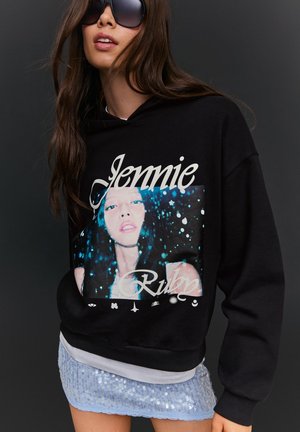 Donna che indossa una felpa nera oversize con la scritta "Jennie Ruby" e la grafica del volto di una donna, abbinata a una gonna blu con paillettes.