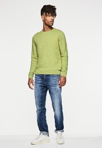 Suéter verde de cuello redondo, tejido con textura; jeans de mezclilla azul; zapatillas blancas con detalles en verde; pose de pie frente a un fondo blanco.