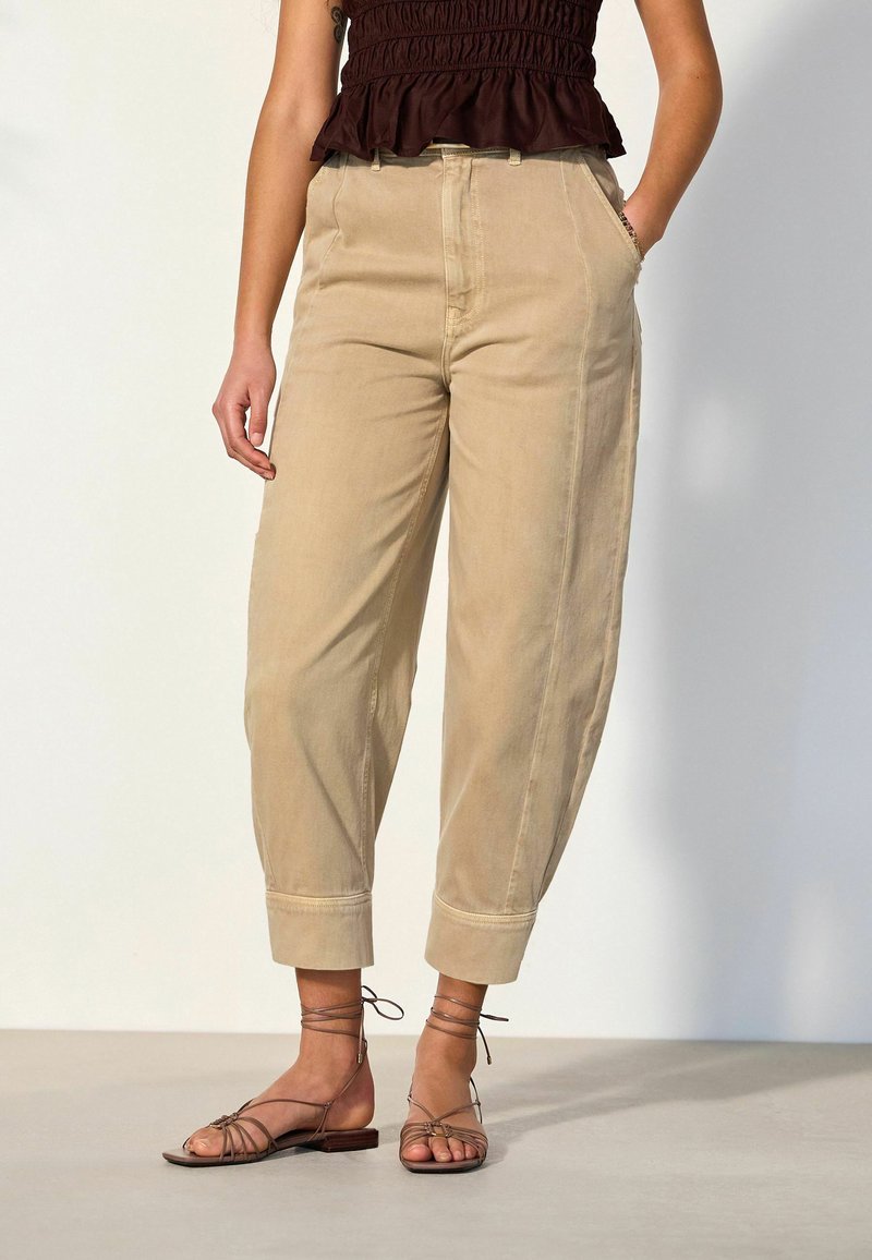 Woman wearing beige cuffed pants, dark brown sleeveless top, and brown strappy flat sandals, standing with one hand in pocket.