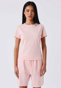 Camiseta de algodón de color rosa claro con mangas cortas y cuello redondo, que presenta un logo bordado en blanco en el pecho. Textura suave, ajuste casual.