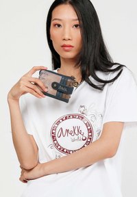 Una donna con lunghi capelli neri tiene un piccolo portafoglio multicolore zip vicino al petto, indossando una maglietta bianca con un logo circolare rosso "Anekke World".