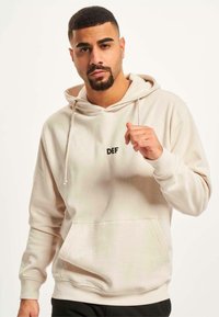 Lys beige hettegenser laget av myk stoff, med en frontlomme og svart "DEF"-logo på brystet, samt snorer på hetten.