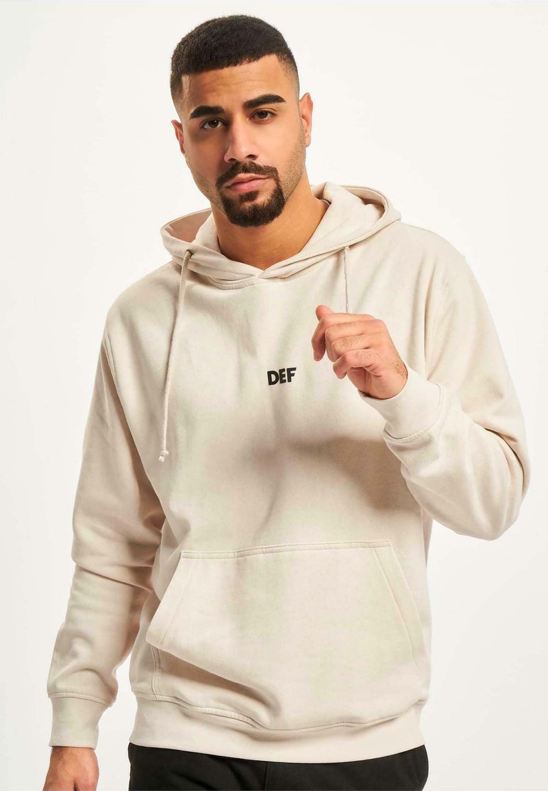 Lys beige hettegenser laget av myk stoff, med en frontlomme og svart "DEF"-logo på brystet, samt snorer på hetten.