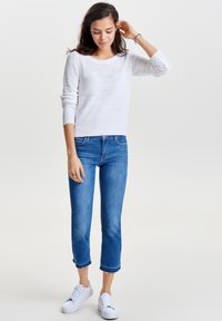 Maglione bianco lavorato a maglia, jeans corti blu con orlo sfrangiato e sneakers bianche. Le texture includono una maglia larga e tessuto di denim.