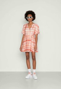 Vestido xadrez rosa com mangas bufantes curtas, decote em V profundo e cintura com laço. Combinado com ténis brancos de plataforma e óculos de sol.