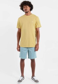 T-shirt en coton jaune à col rond, associé à un short bleu clair. Le mannequin porte des baskets grises avec des lacets blancs, debout sur un fond uni.
