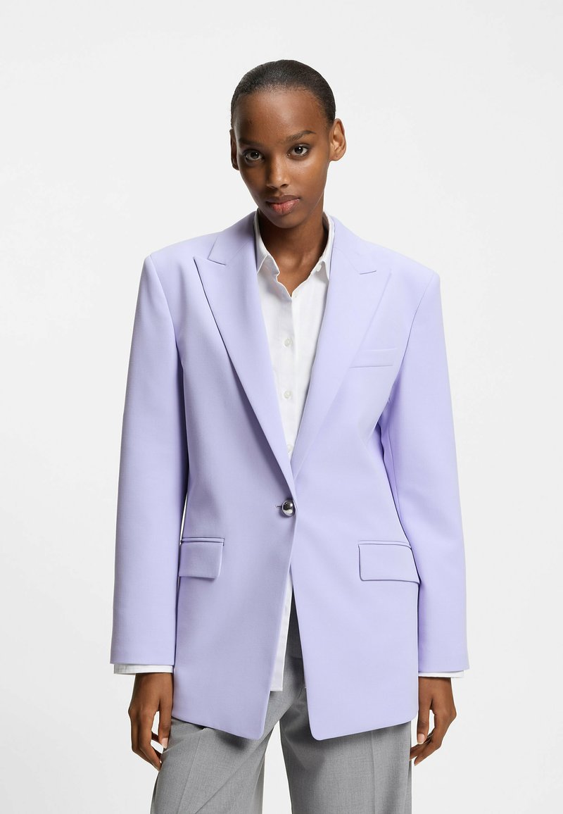 Veste violet clair à la coupe structurée, fermeture à un bouton, revers crantés et deux poches à l'avant, portée sur une chemise blanche.
