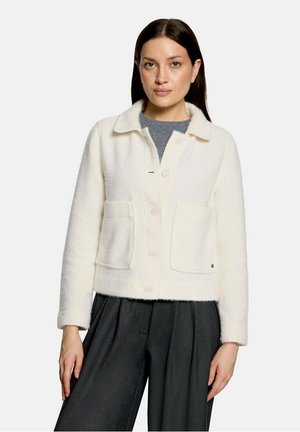 Femme aux cheveux raides et foncés portant une veste blanche boutonnée avec de grandes poches et un pantalon large sombre, debout devant un fond uni.