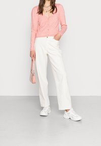 Rosa stickad cardigan med långa ärmar och knappar, ihop med vita vida byxor och vita sportskor. Accessoar: liten rosa handväska.