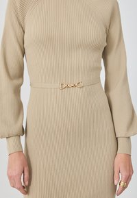 Beige ribbet kjole med lange ærmer, prydet med et bælte med guldhardwarebuckle i taljen. Blød tekstur, tætsiddende silhuet.