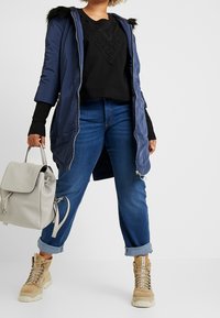 Granatowa pikowana kurtka z kapturem z sztucznego futra, czarny top z koronkowym zdobieniem, niebieskie dżinsy i beżowe masywne buty, trzymająca jasnoszary plecak.