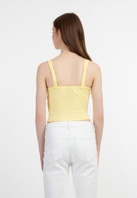 Keltainen raidoitettu crop-top, jossa on leveät hihnat, napakasti istuva muotoilu ja sileä pinta. Yhdistetty valkoisiin korkeavyötäröisiin farkkuihin, näkyy takaapäin.