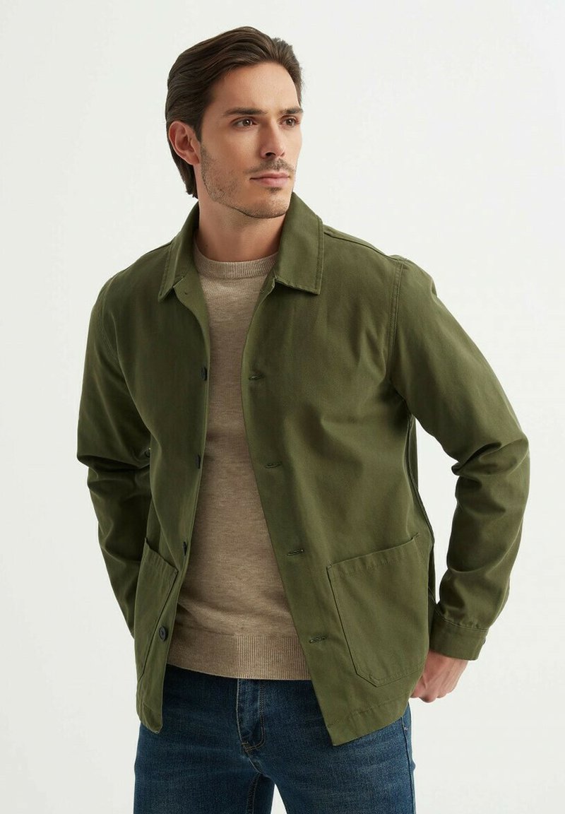 Veste vert olive avec un devant boutonné et deux poches latérales, portée sur un pull beige. Présente un col et une texture lisse.