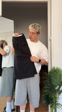 Pantaloni neri con pieghe, dettagli della vita e una trama liscia indossati da una persona che indossa una camicia bianca e pantaloni corti grigi.