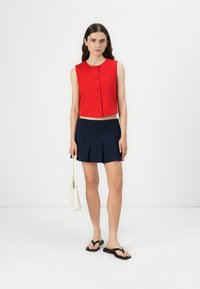 Chaleco rojo recortado con cinco botones, combinado con una falda plisada azul marino. Los accesorios incluyen un bolso blanco y chanclas negras.