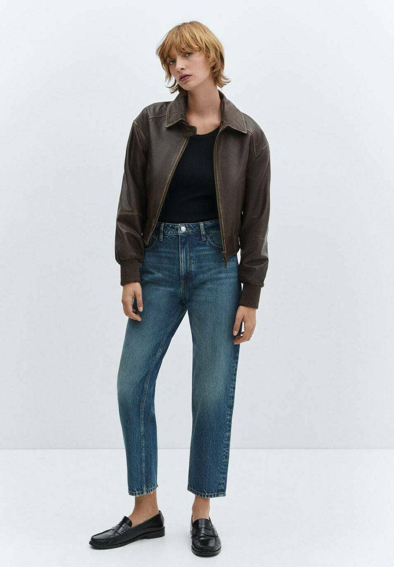 Braune Lederbomberjacke, lange Ärmel mit gerippten Bündchen, kombiniert mit hochgeschnittenen blauen Denim-Jeans und schwarzen Slippern.